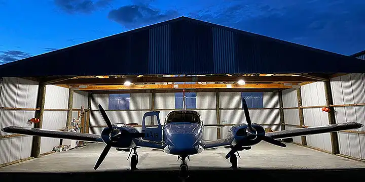 Golden Bay Airs twin engine aircraft parked in front of hangar in the evening, lights are on so the aircraft is silhouetted