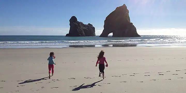 Two children run on the sand towards the Whaririki Sea Arches that are silhouetted against a blue sky