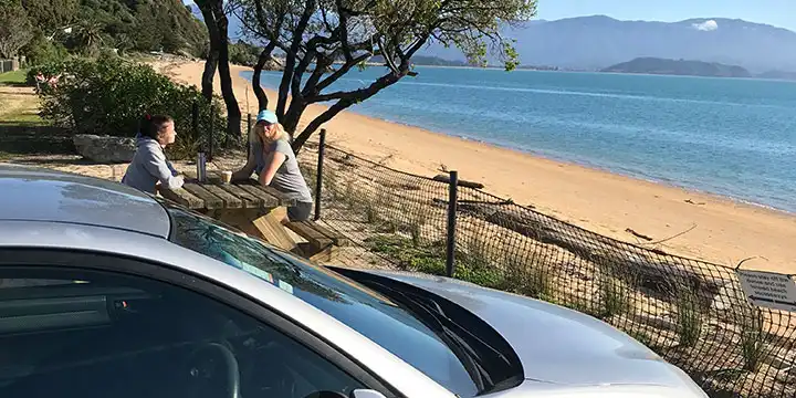 Two people sit on a picnic table at Tata Beach in Golden Bay with their rental car in the foreground, golden sand beach in the background