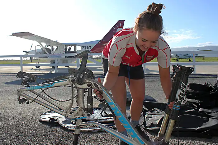 A biker smiling as she packs up her bike at Karamea Aerodrome, Golden Bay Air Airvan in the background