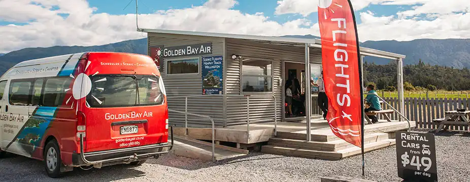 Golden Bay Air van parked outside the Golden Bay Air terminal at Takaka Airport. People sit on the deck enjoying the sun
